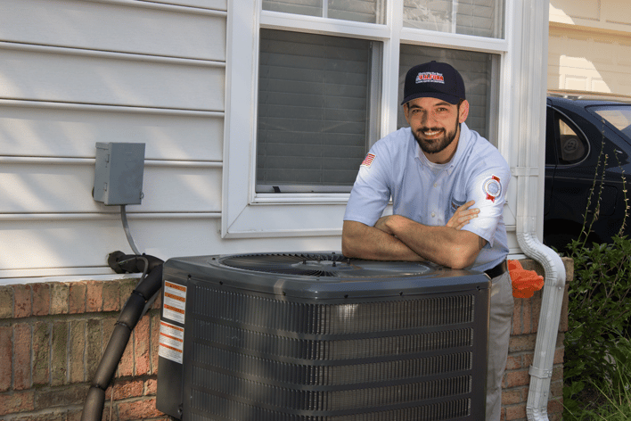 Technician smiling and leaning forearms on an outdoor HVAC unit next to a white home.